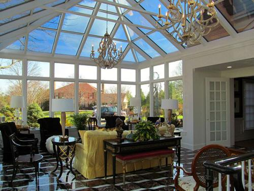 : A conservatory interior with tile floors, chandeliers, sitting room furniture, and windows all along the roof and back wall.